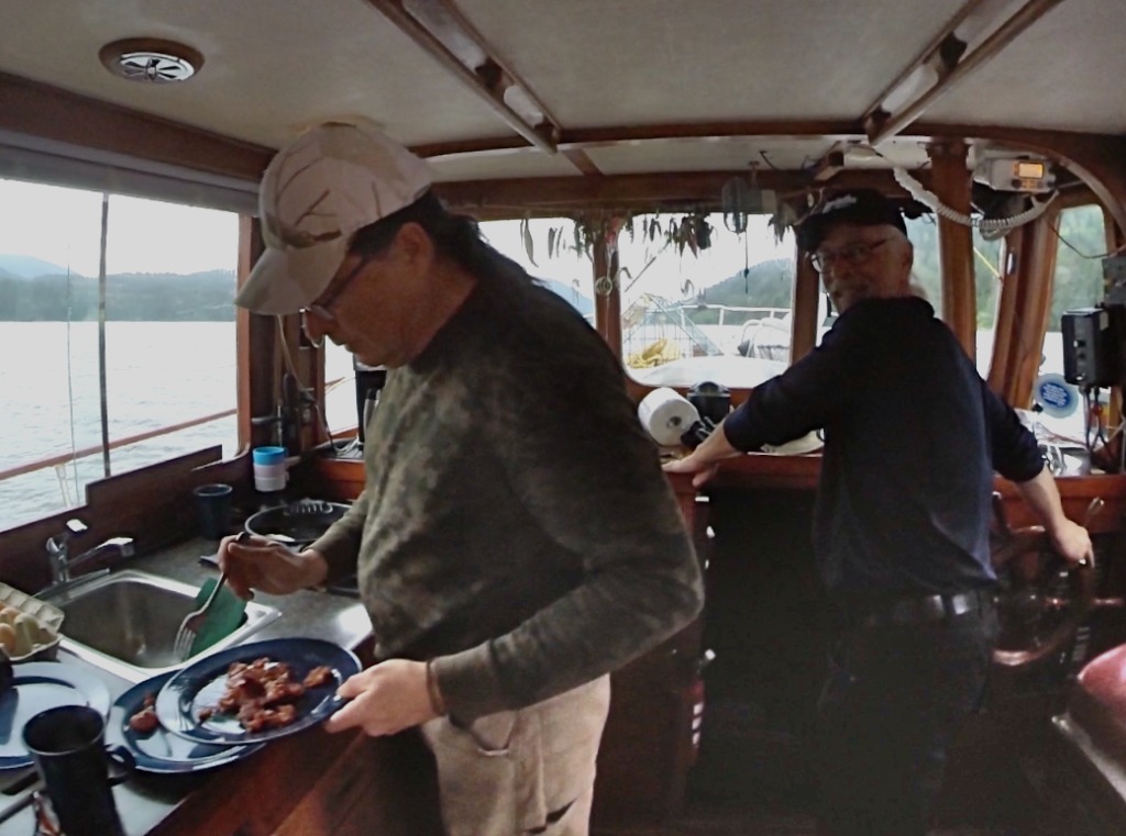 man driving the boat while other one makes breakfast